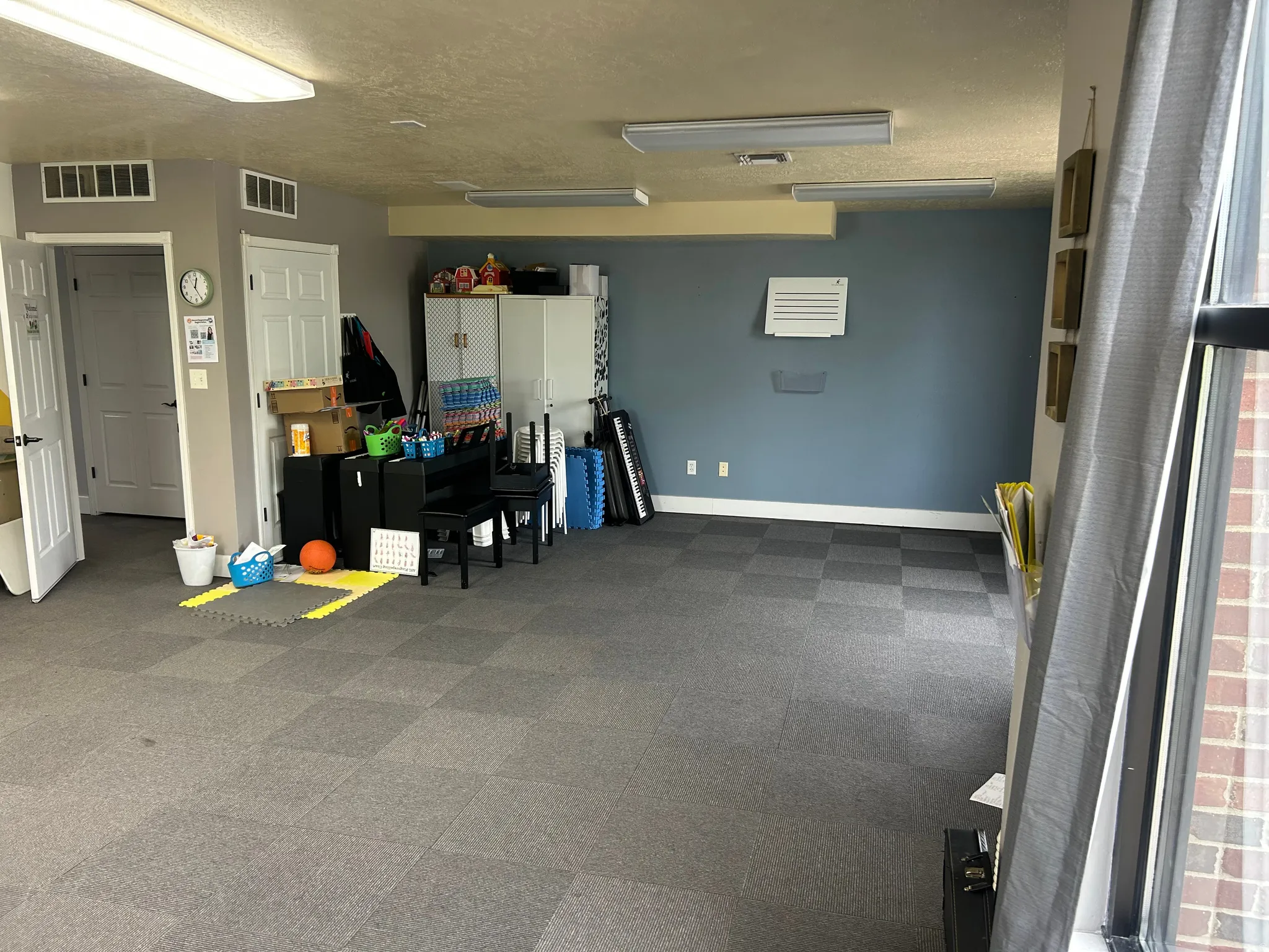 Spacious room featuring gray modular carpet tiles, two white paneled doors, exposed ceiling beams, and fluorescent lighting fixtures