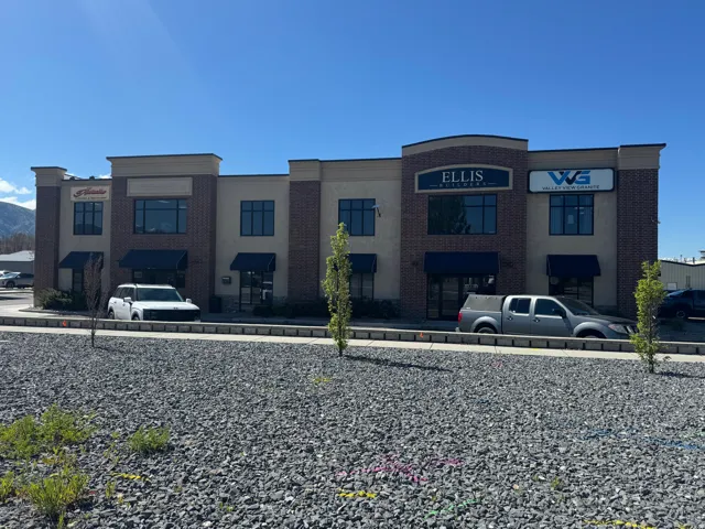 Two-story commercial building featuring brick and stucco facade, arched entryway, multiple window awnings, and an adjacent gravel parking area
