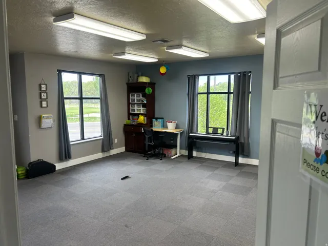 Interior space featuring checkerboard-patterned carpet tile flooring, two large windows with dark grey curtains, and two ceiling-mounted rectangular fluorescent light fixtures