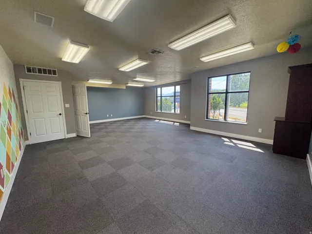Spacious room featuring checkered carpet tile flooring, multiple windows providing natural light, and ample overhead fluorescent lighting