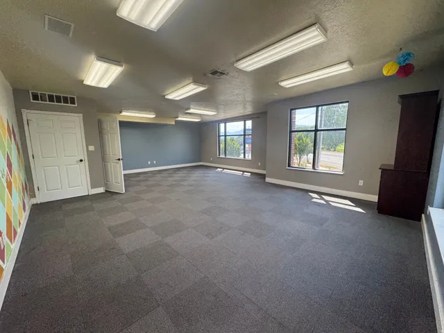 Spacious room featuring checkered carpet tile flooring, multiple windows, and extensive fluorescent ceiling lighting