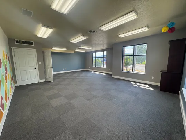 Spacious room featuring checkered carpet tile flooring, multiple windows, and overhead fluorescent lighting