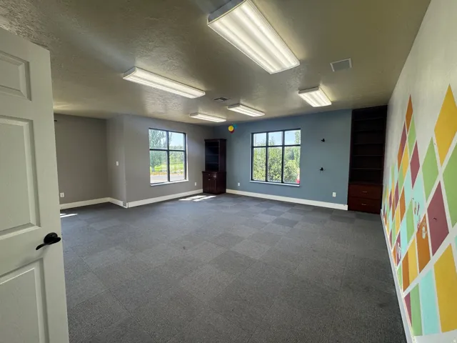 Spacious room featuring modular carpet flooring, multiple windows, a built-in shelving unit, and overhead fluorescent lighting