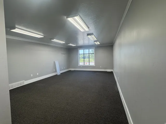 Spacious interior room featuring grey wall paint, dark patterned carpeting, recessed baseboard heating, and ample natural light from a multi-pane window