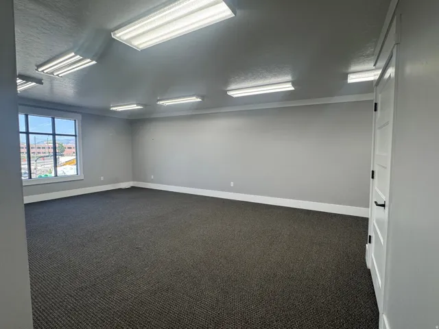 Expansive carpeted room featuring multiple overhead fluorescent light fixtures, a large window with white trim, and a white paneled door with black hardware