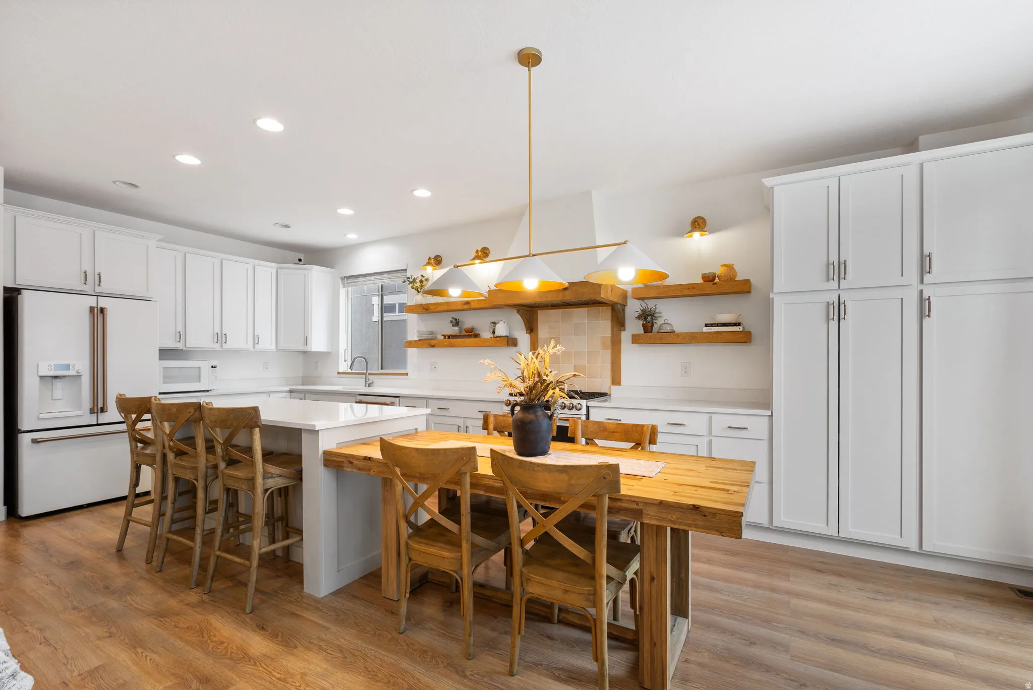 Open-concept kitchen featuring white shaker-style cabinetry, a central island with countertop seating, stainless steel appliances, wood-finish flooring, and open wood shelving