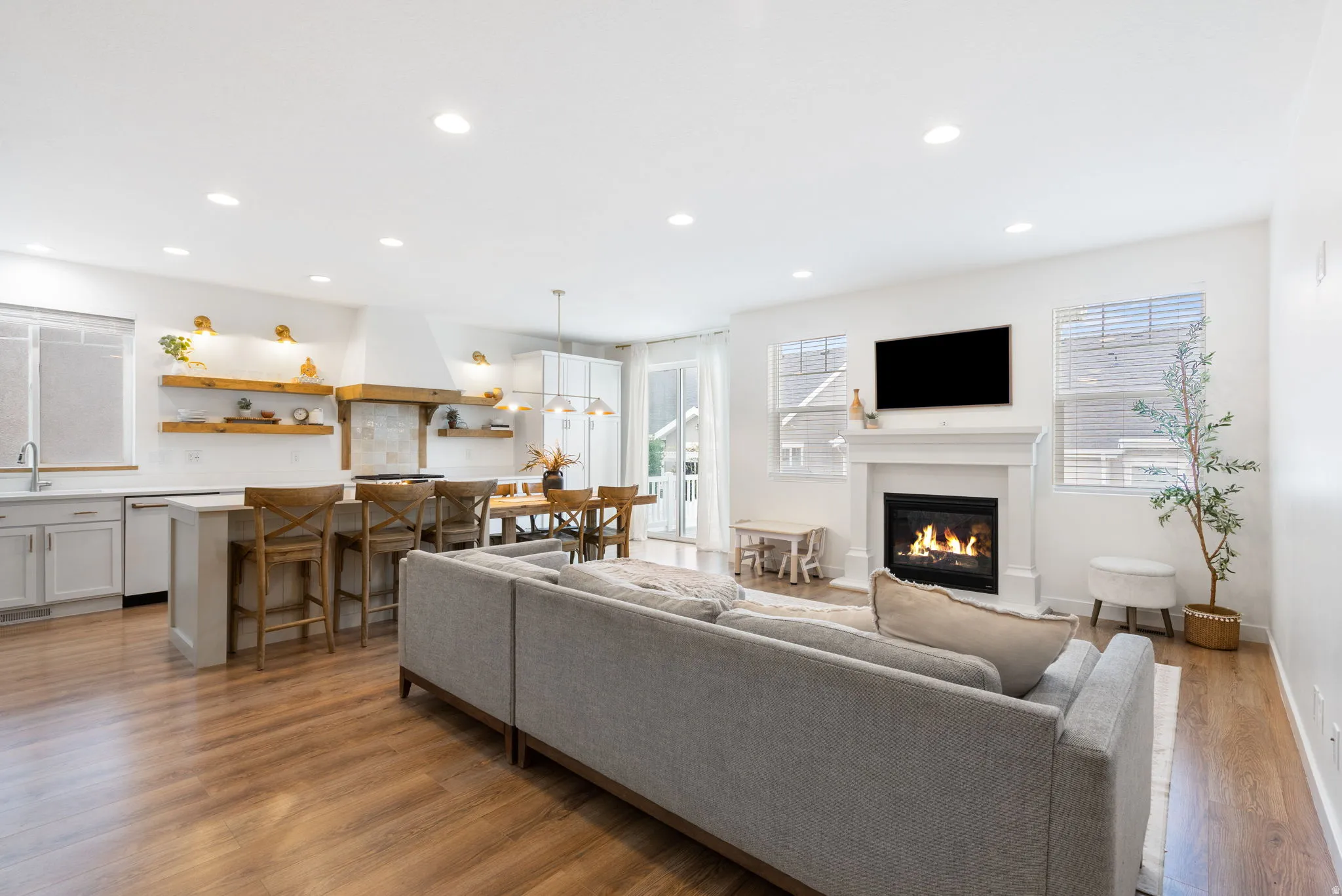 Open-concept living space featuring wood-finish flooring, a white mantel fireplace, recessed lighting, a kitchen island with seating, and a range hood with open shelving