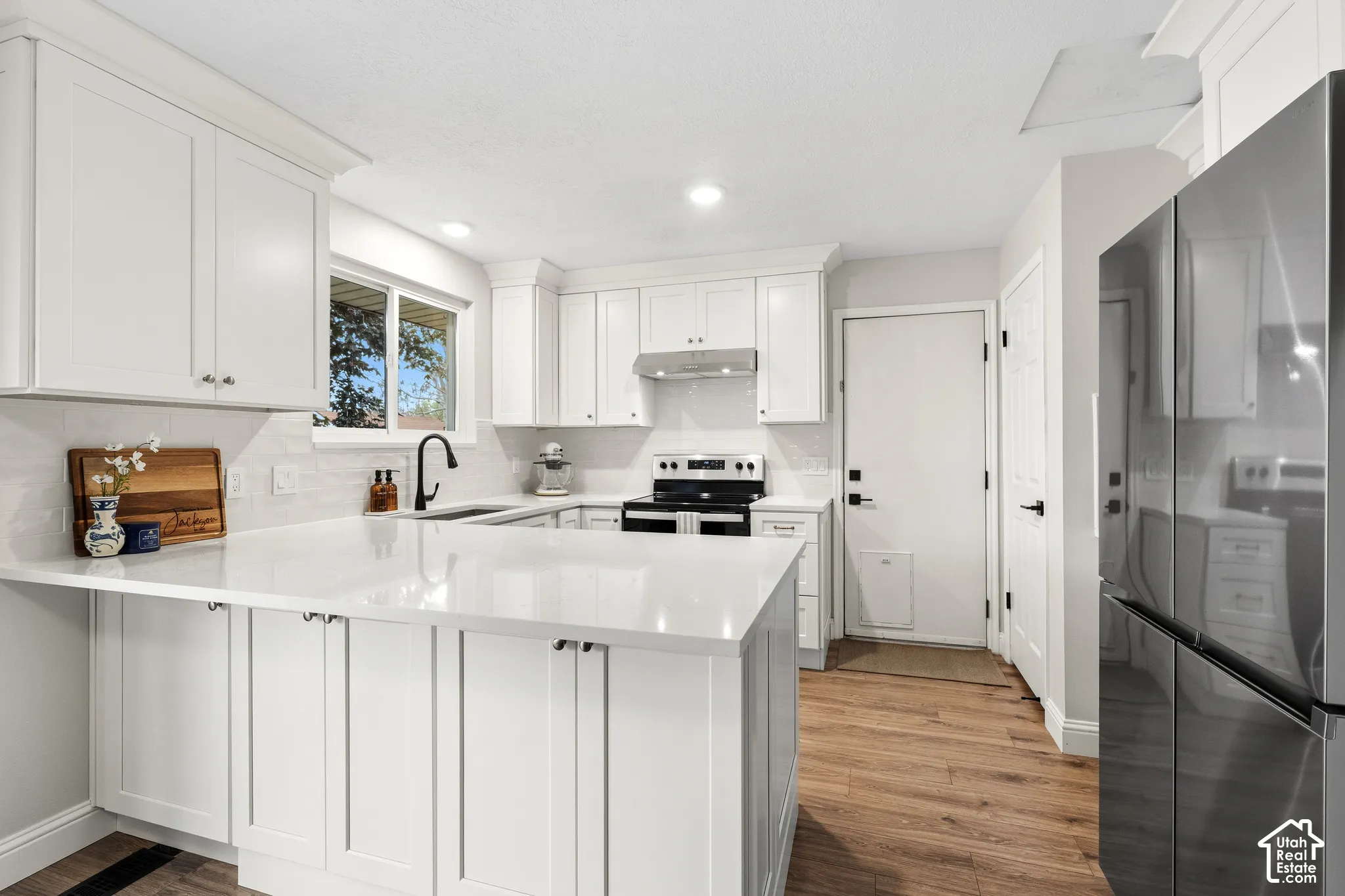 Modern kitchen featuring white shaker cabinetry, light-toned countertops, a stainless steel range with overhead hood, and a black matte faucet