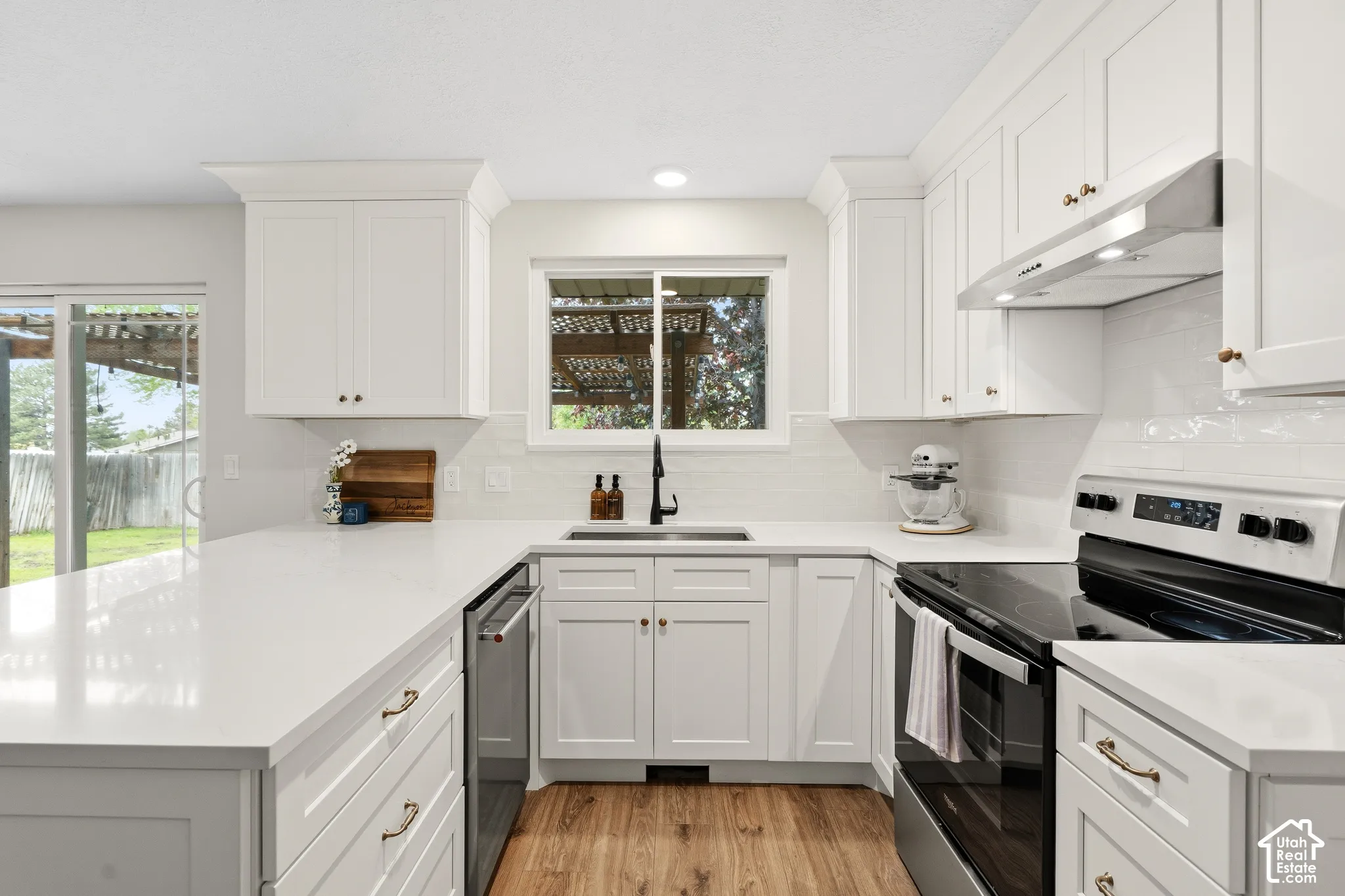 Contemporary kitchen featuring white shaker cabinetry, light-toned countertops, stainless steel appliances, wood-finish flooring, and a sliding glass door to the exterior