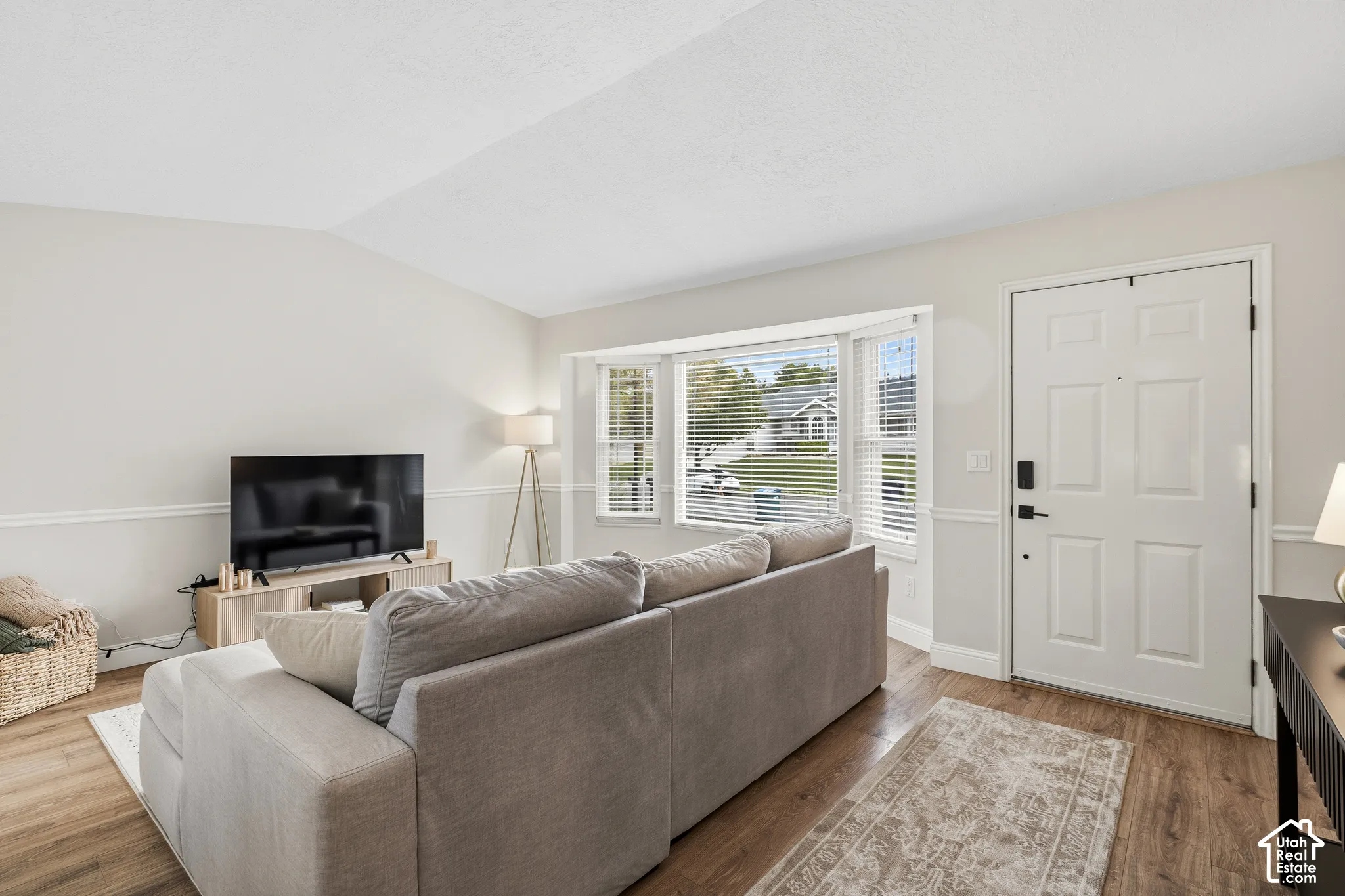 Living area featuring wood-finish flooring, a white paneled front entry door, and a bay window with horizontal blinds