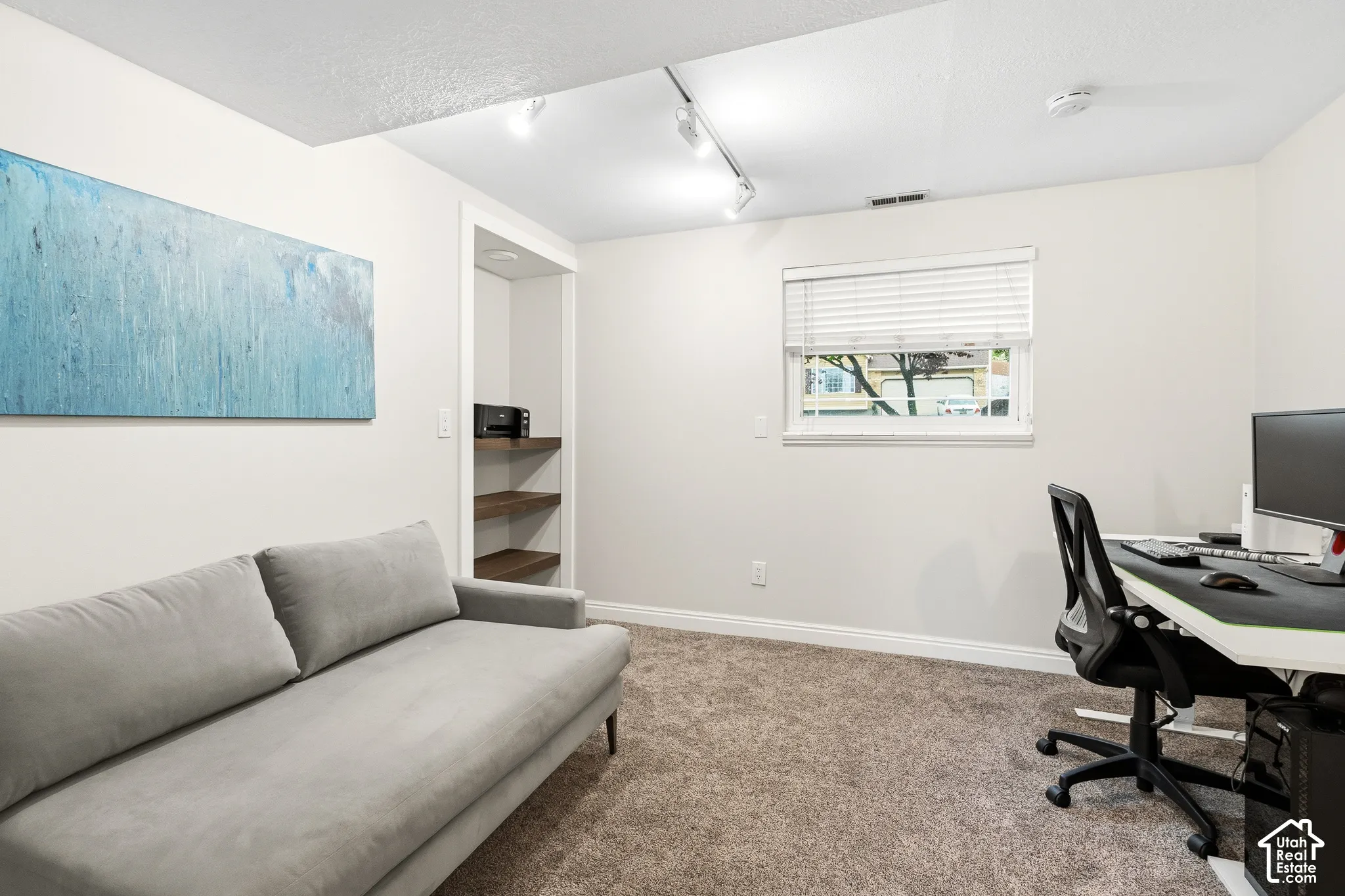Carpeted interior space featuring a window with blinds, track lighting, and a built-in shelving unit with wood-finish shelves