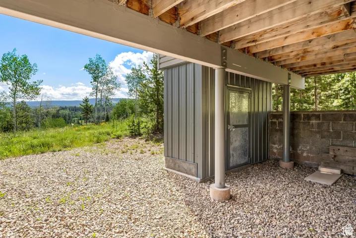 Gravel outdoor area featuring a corrugated metal storage shed, exposed wood deck joists, steel support beams, and a concrete block retaining wall