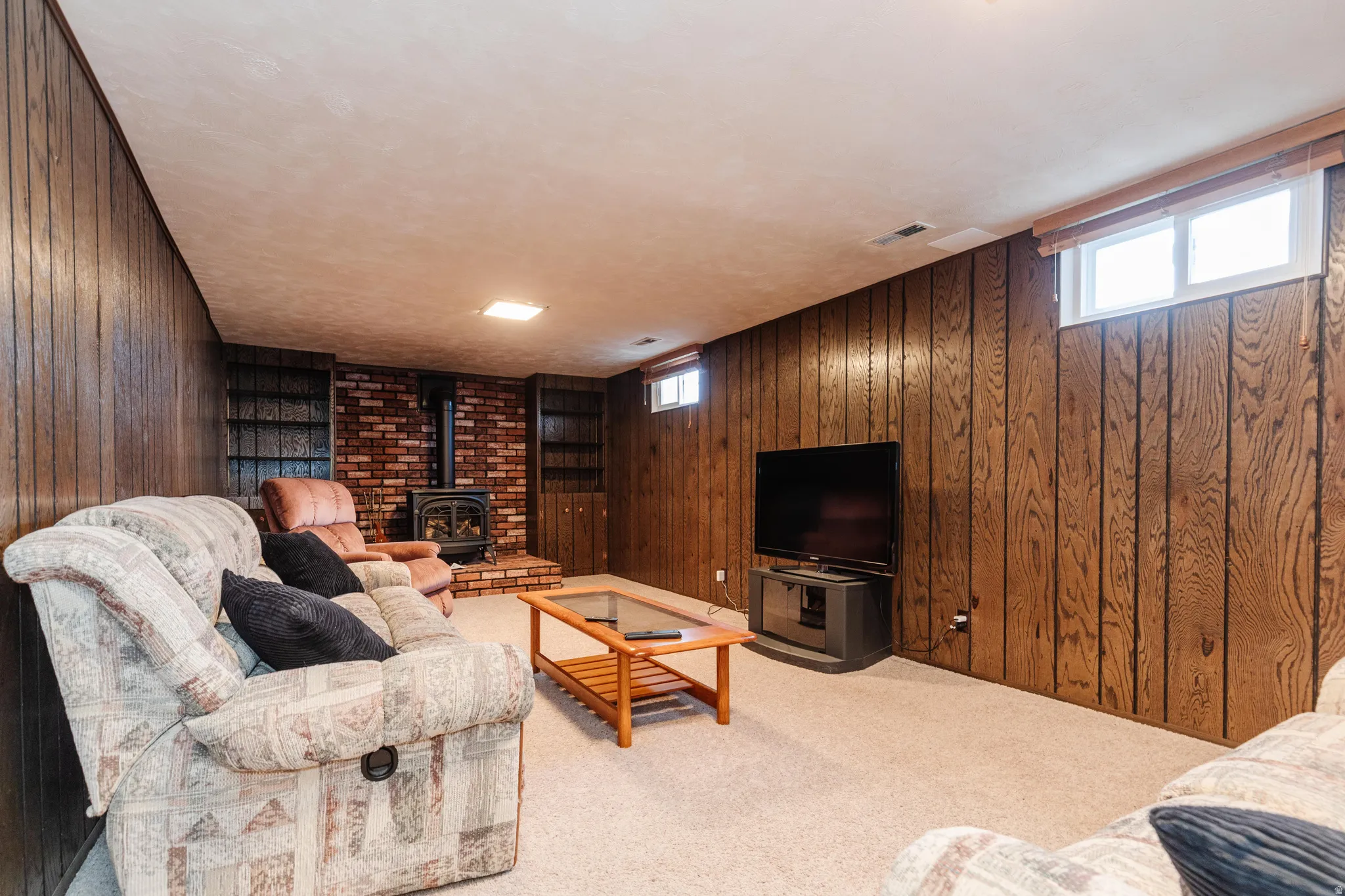 Carpeted recreation room featuring wood paneling, a brick-faced wood-burning stove, built-in shelving, and two windows for natural light
