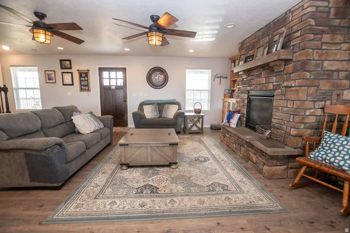 Spacious living area featuring a stone fireplace with a wood mantle, wood-finish flooring, two ceiling fans with integrated lighting, and multiple windows for natural light