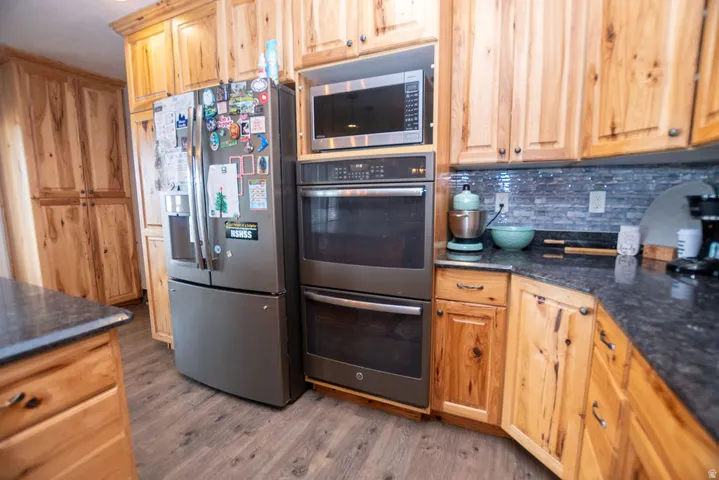 Kitchen featuring natural wood cabinetry, dark speckled countertops, stainless steel appliances including a built-in double oven and microwave, and a tiled backsplash