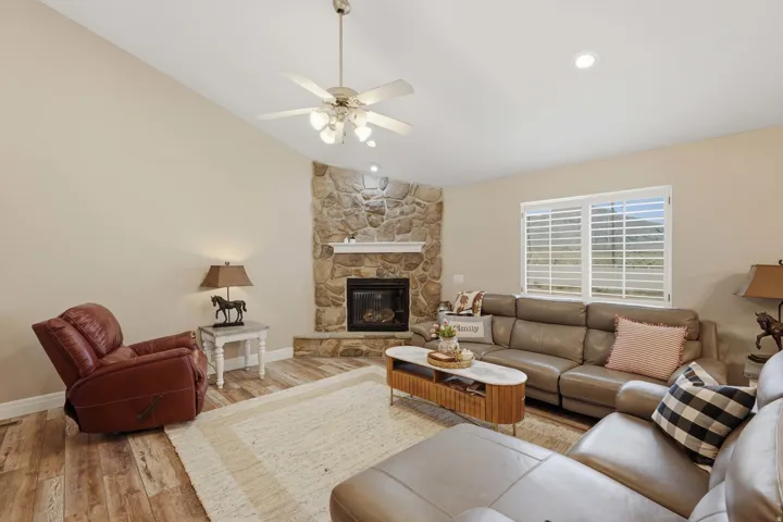 Corner stone fireplace with a white mantel, vaulted ceiling, wood-finish flooring, ceiling fan, and window with plantation shutters