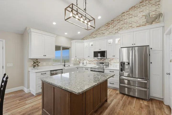 Bright kitchen featuring a granite-topped island, white cabinetry, stainless steel appliances, wood-finish flooring, and a vaulted ceiling with a brick accent wall