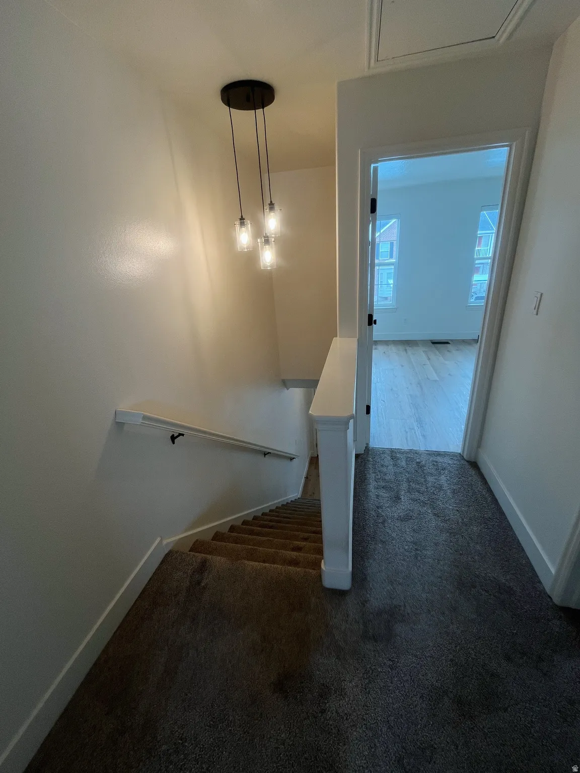 Staircase with dark carpet and white railing featuring a modern pendant light fixture and access to a room with wood-finish flooring
