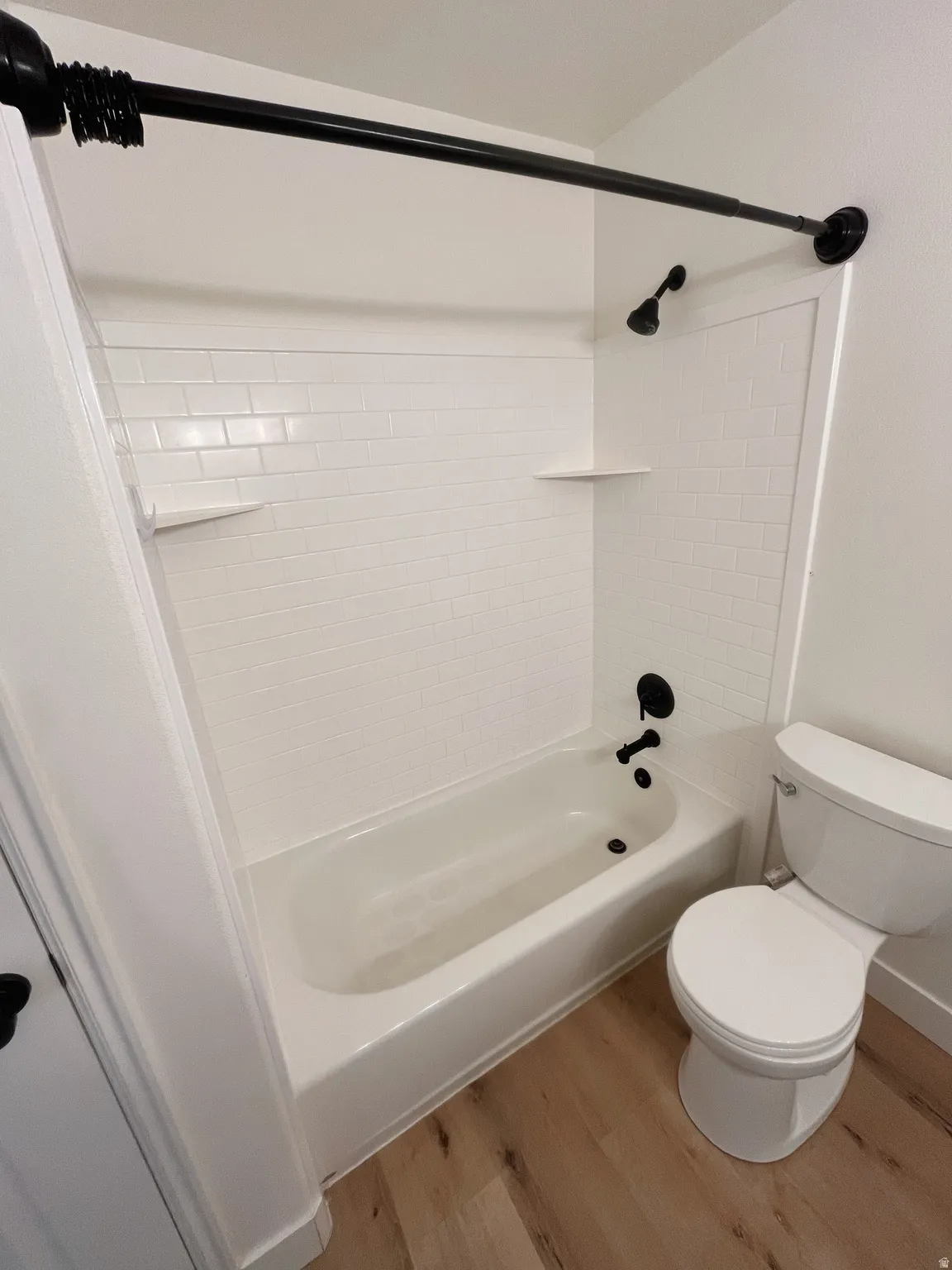 White subway tile shower surround with integrated shelving and matte black fixtures, paired with a white bathtub and toilet, set on wood-finish flooring