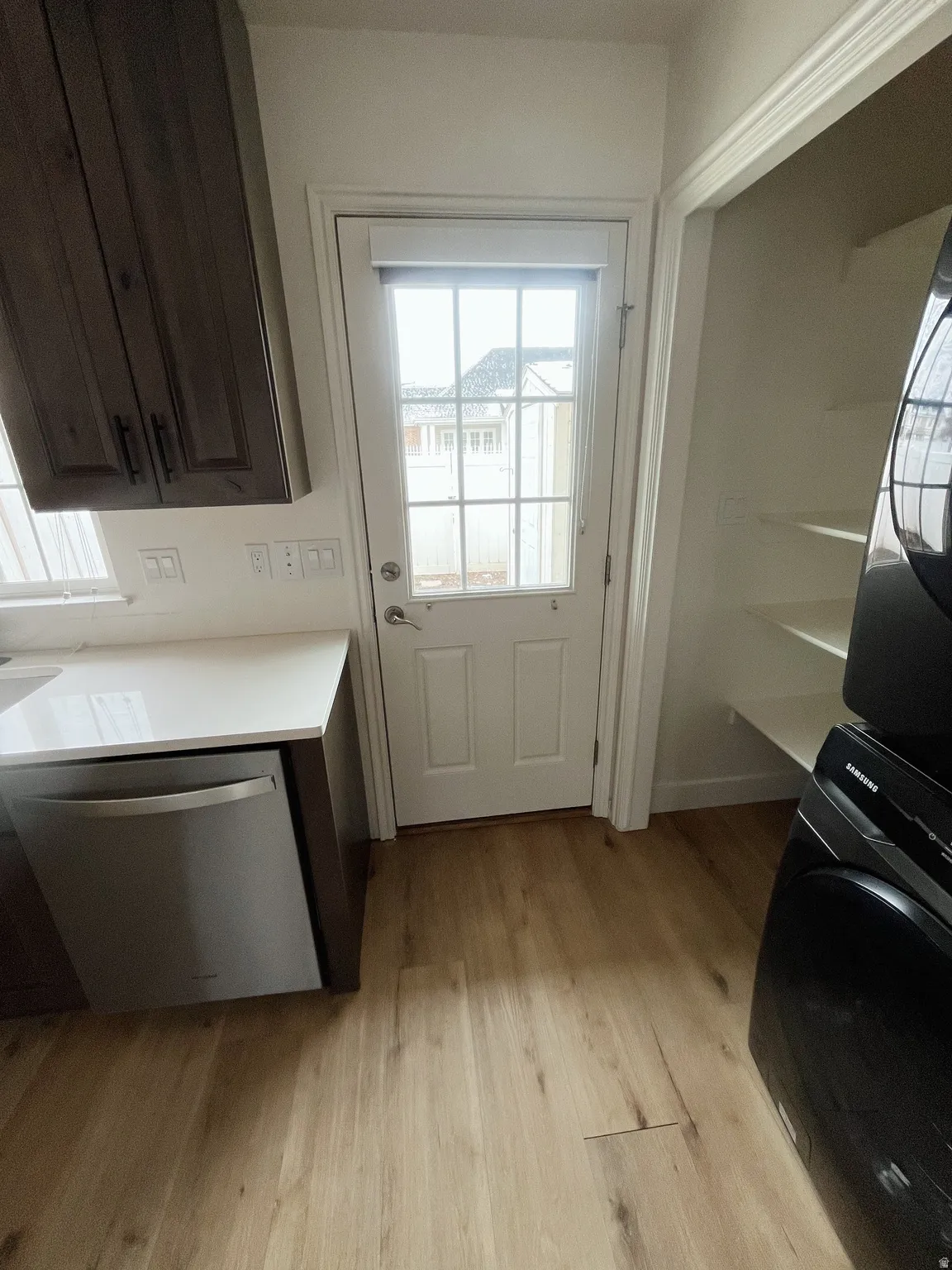 Wood-finish flooring throughout, featuring a full-light paneled exterior door with blinds, dark wood-finish cabinetry, and a built-in stainless steel dishwasher