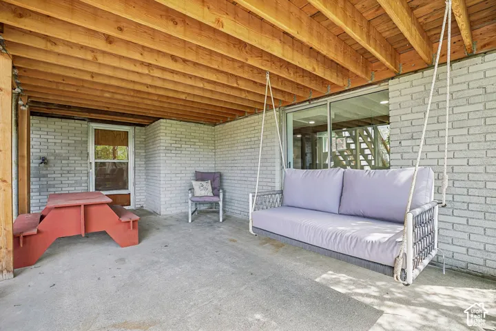 Covered patio space featuring an exposed wood ceiling, light-colored brick walls, concrete flooring, and a glass sliding door
