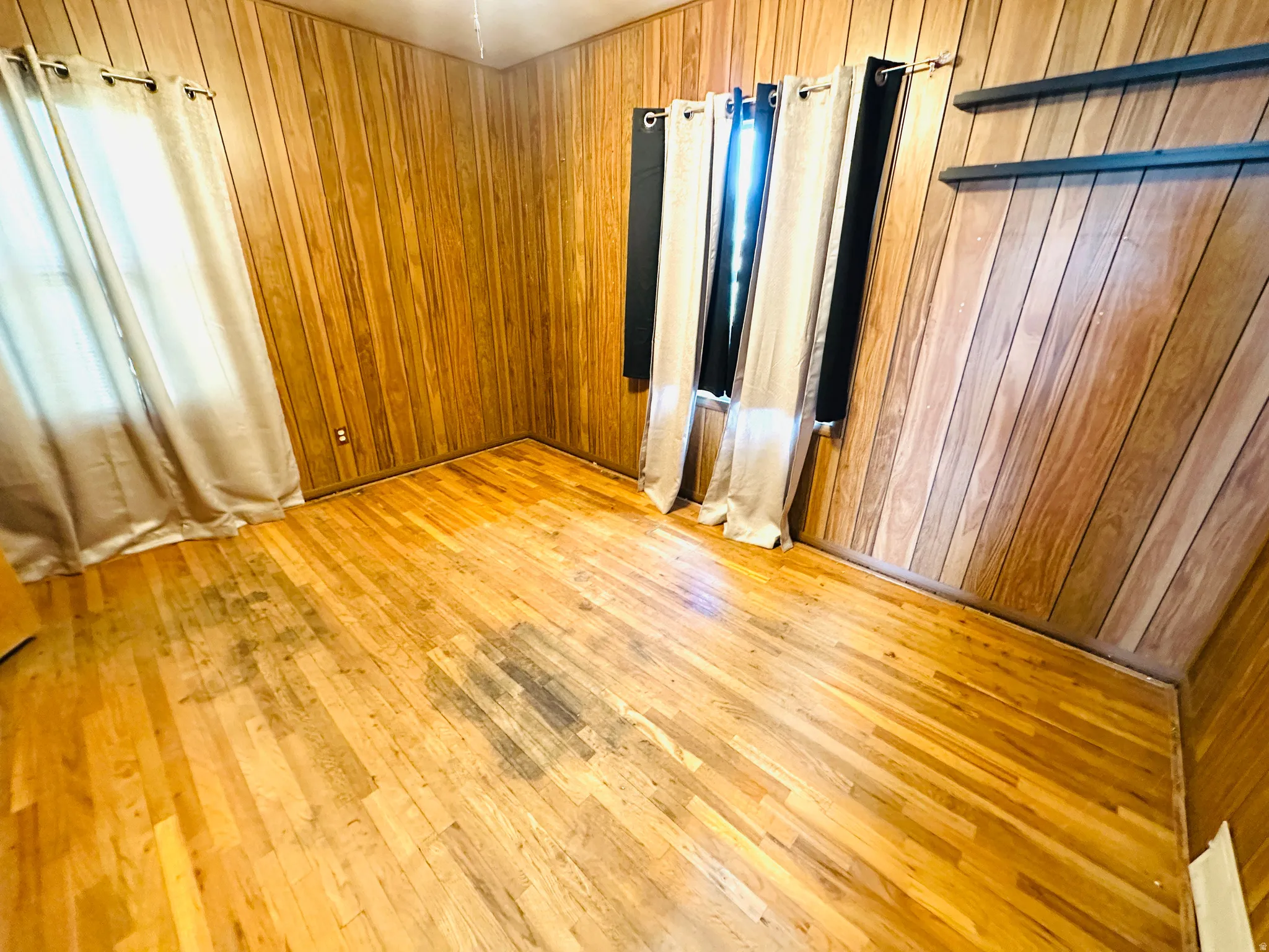 Wood-paneled room featuring light wood-finish flooring, two windows, and a wall-mounted shelf