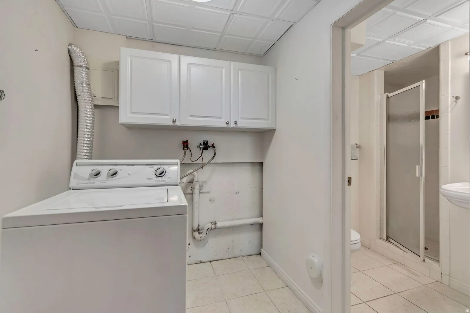 Dedicated laundry area featuring white upper cabinetry, tiled flooring, and an adjacent three-piece bath with a glass-enclosed shower