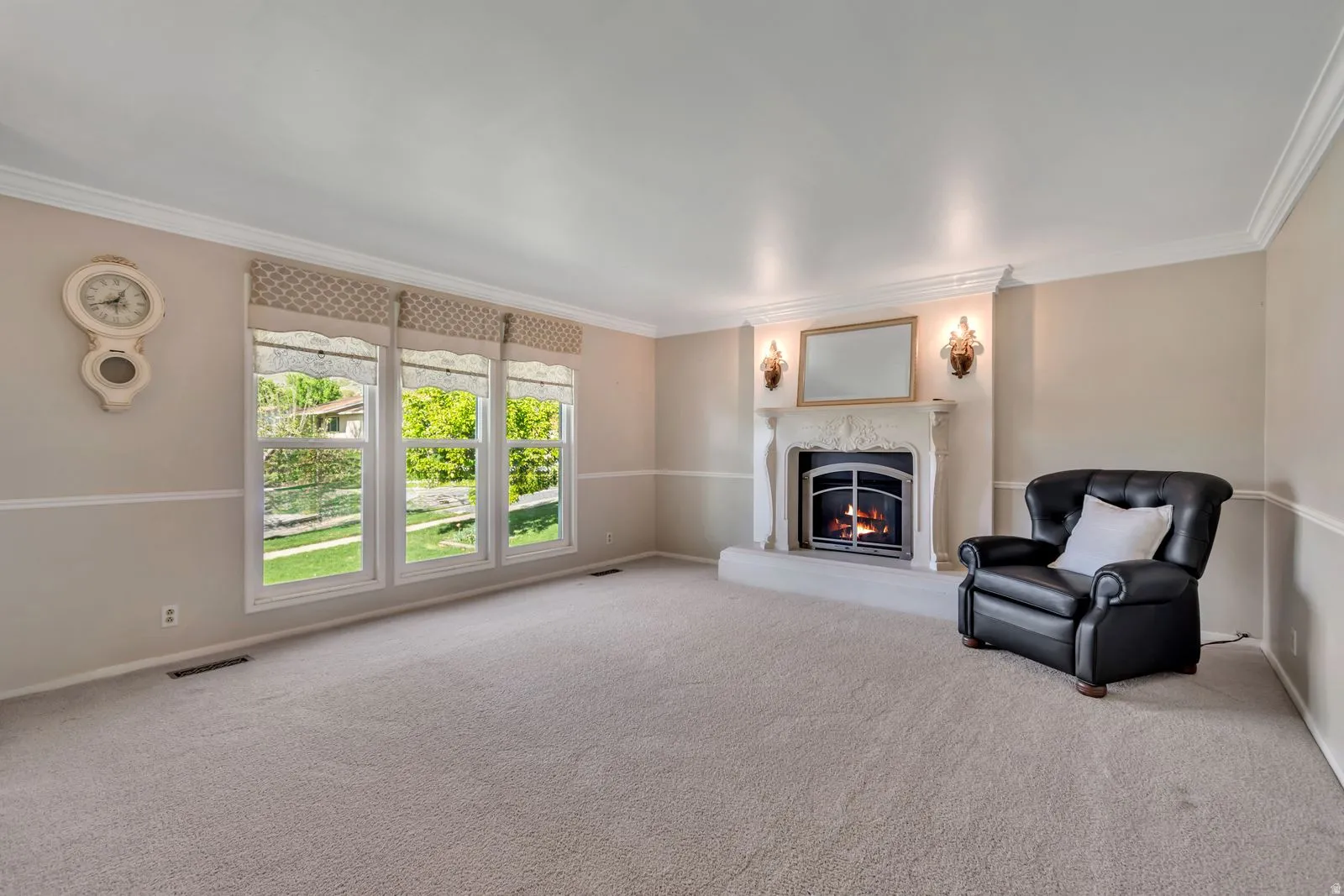 Living area featuring a detailed fireplace with an ornate mantel, crown molding, chair rail molding, three large windows, and wall sconces