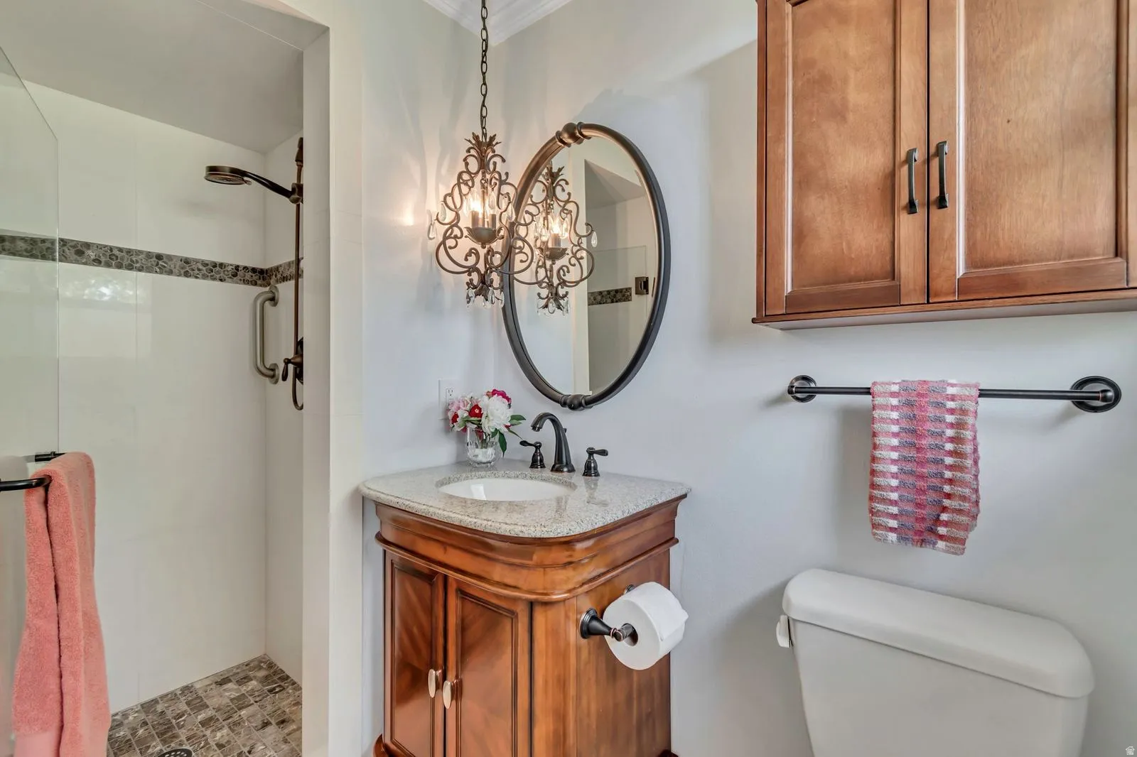 Bathroom featuring a freestanding wood vanity with a white countertop and undermount sink, an ornate hanging chandelier, a frameless glass shower with a tiled floor and accent band, and a wall-mounted wood cabinet