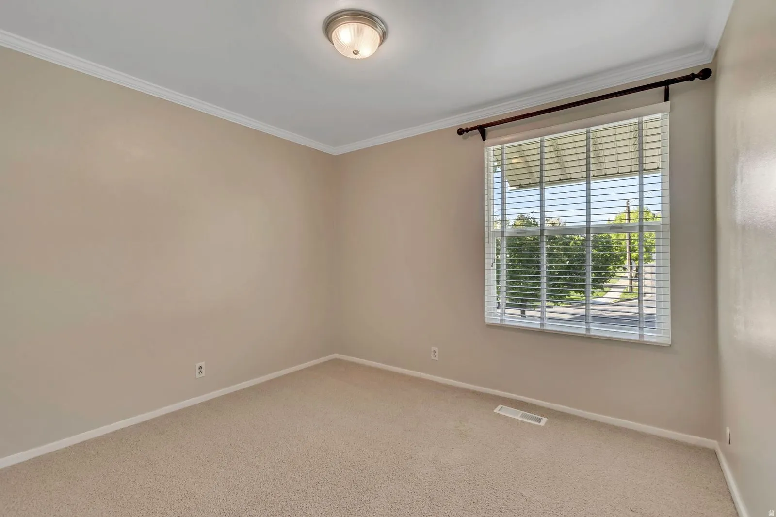 Neutral-toned room featuring a single window with horizontal blinds, tan carpet, beige walls, white crown molding, and a flush-mount ceiling light