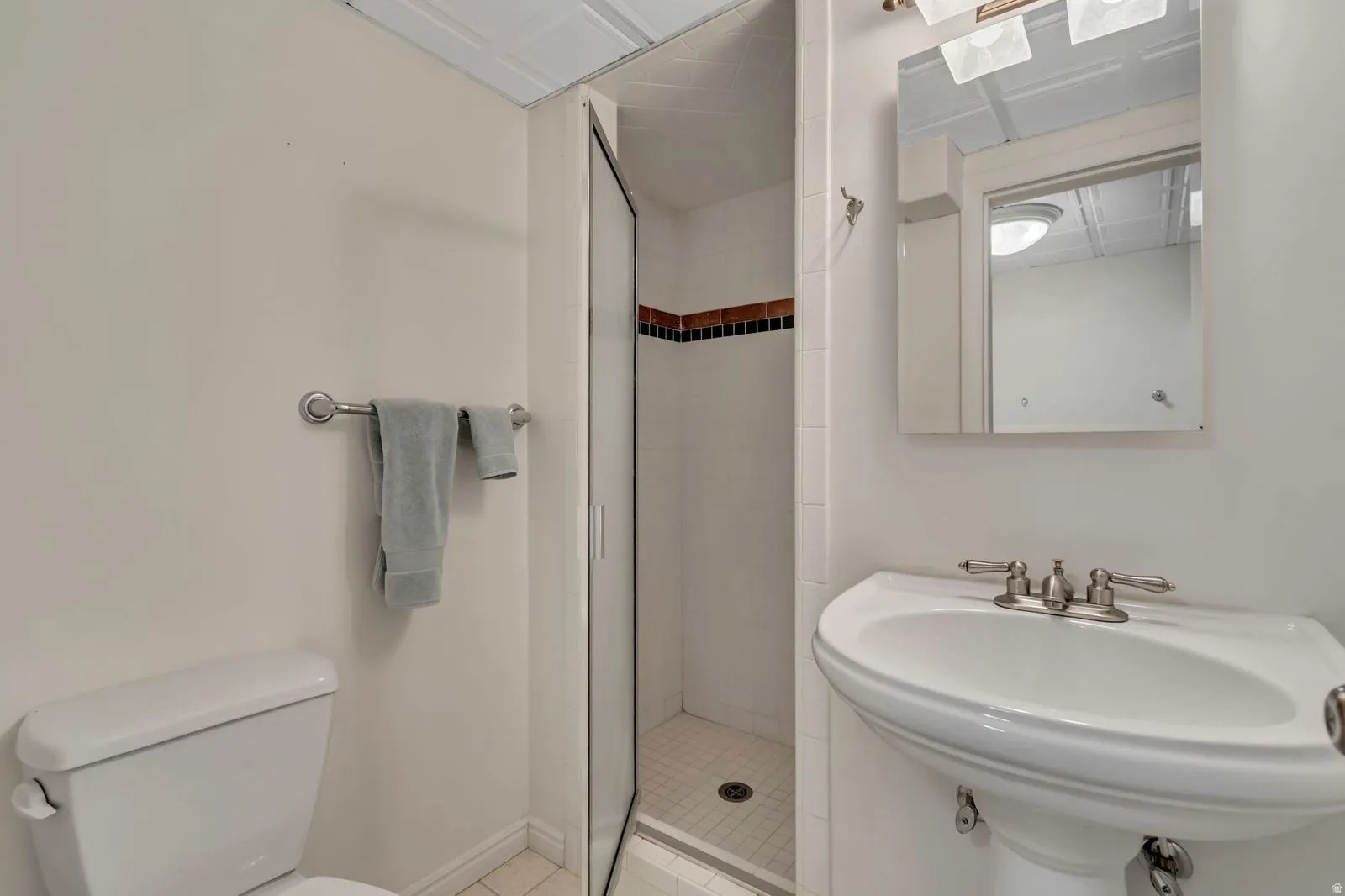 Pedestal sink with brushed nickel hardware, mirrored medicine cabinet, glass-enclosed shower with tile accent band, white toilet, and white tile flooring