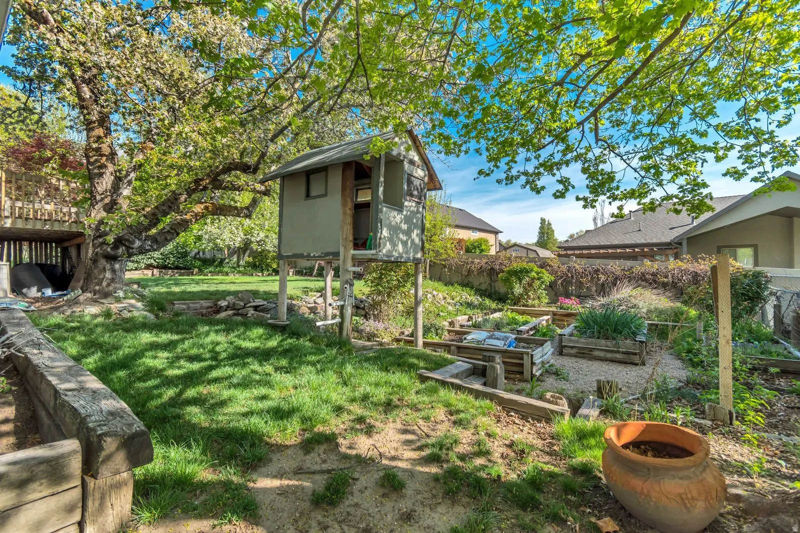 Expansive backyard featuring a manicured lawn, established landscaping, raised garden beds, a built-in playhouse, and mature trees providing natural shade