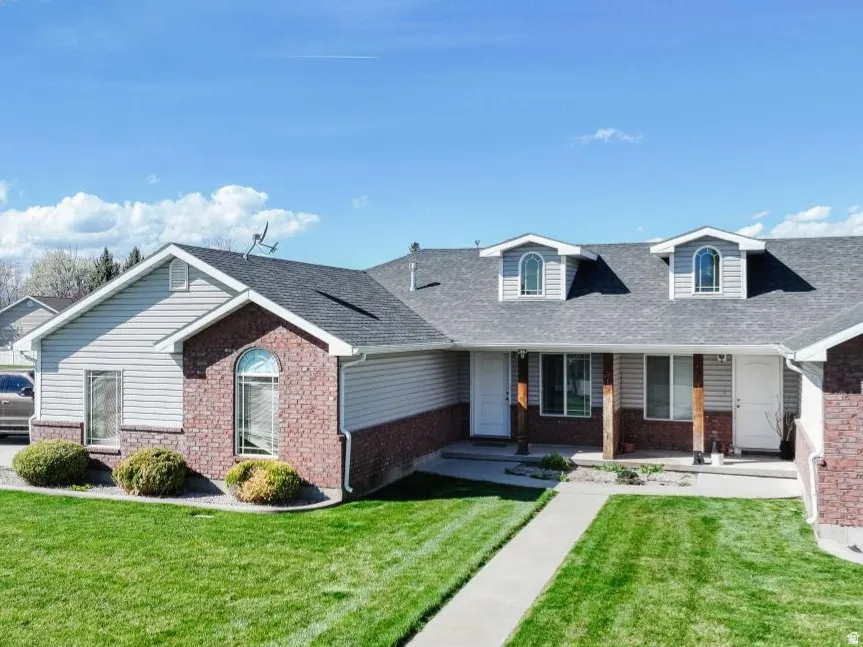 Front exterior featuring gray siding, red brick accents, two dormer windows with arched tops, a covered porch entry with wood-wrapped columns, and a concrete walkway