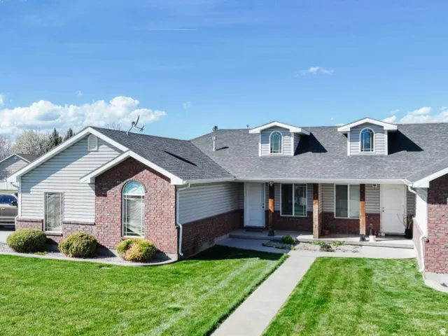 Front exterior featuring gray siding, red brick accents, two dormer windows with arched tops, a covered porch entry with wood-wrapped columns, and a concrete walkway