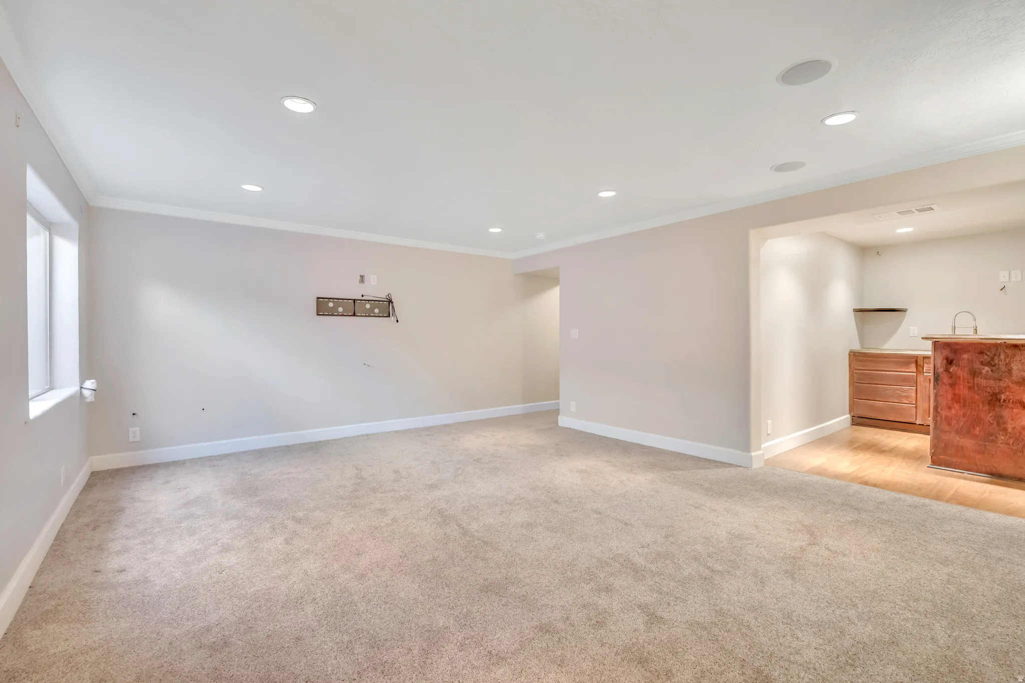 Spacious room featuring neutral carpeting, recessed lighting, crown molding, a window, and a built-in wet bar with wood-finish cabinetry and a sink