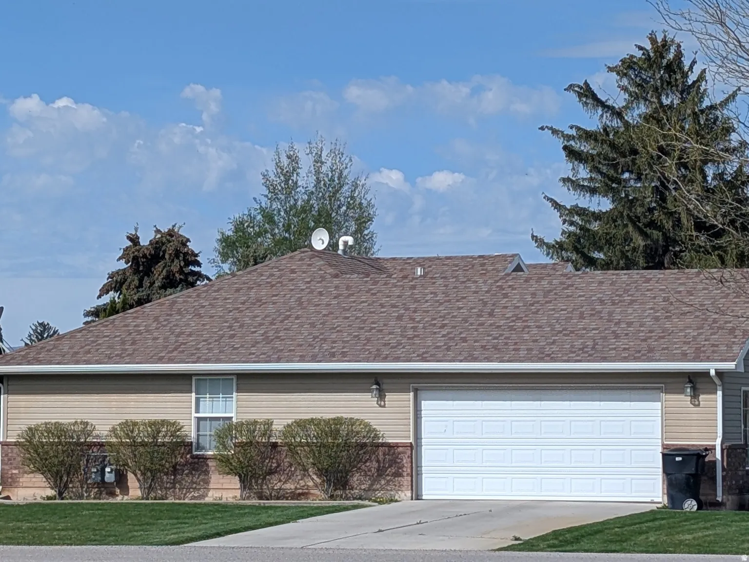 Single-story residence featuring a two-car garage with a paneled door, tan horizontal siding, and a brown asphalt shingle roof
