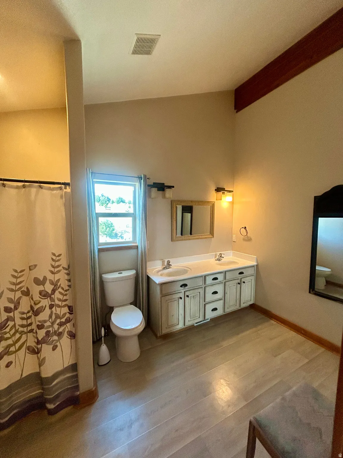 Bathroom featuring a dual-sink vanity with distressed cabinetry, wood-finish flooring, a toilet, and a window