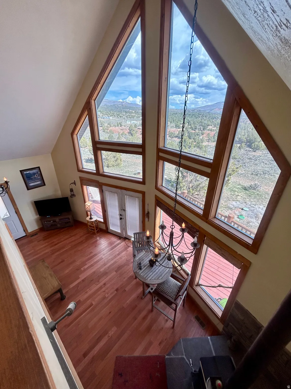 Vaulted ceiling great room featuring expansive A-frame windows, wood-finish flooring, a wrought iron chandelier, and a built-in fireplace with a stone surround