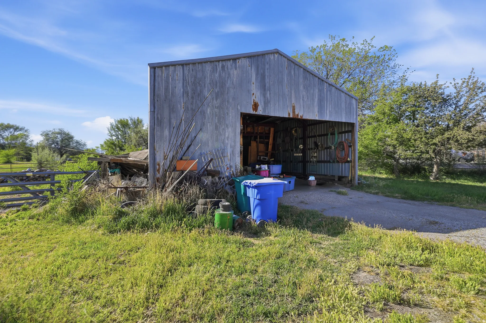 Metal-sided outbuilding with an open bay and gravel driveway, surrounded by a grassy area and mature trees