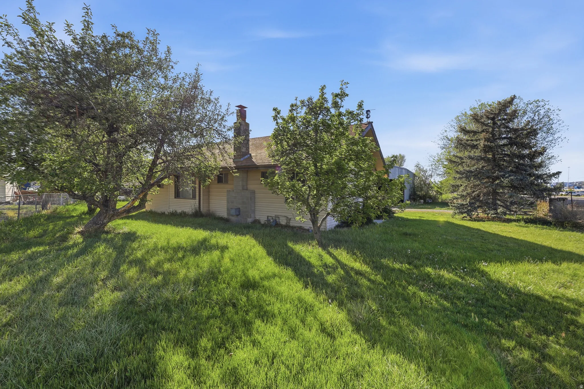 Spacious lawn area with mature trees and a residential structure featuring light-colored siding and a prominent stone chimney