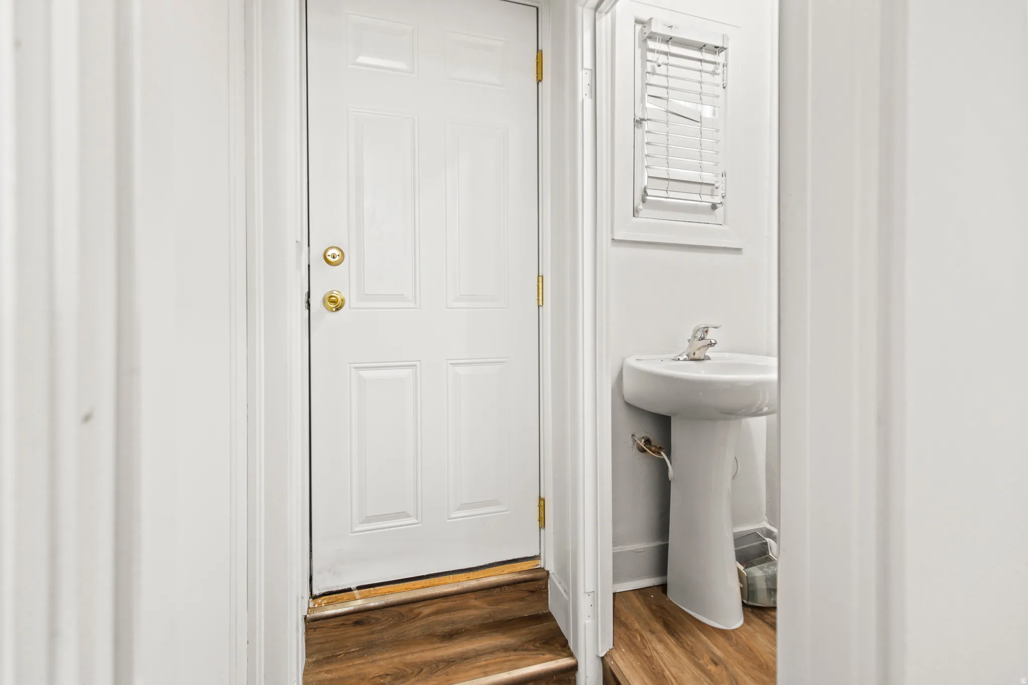 White paneled door with brass hardware, wood-finish flooring, and a white pedestal sink with a window featuring blinds