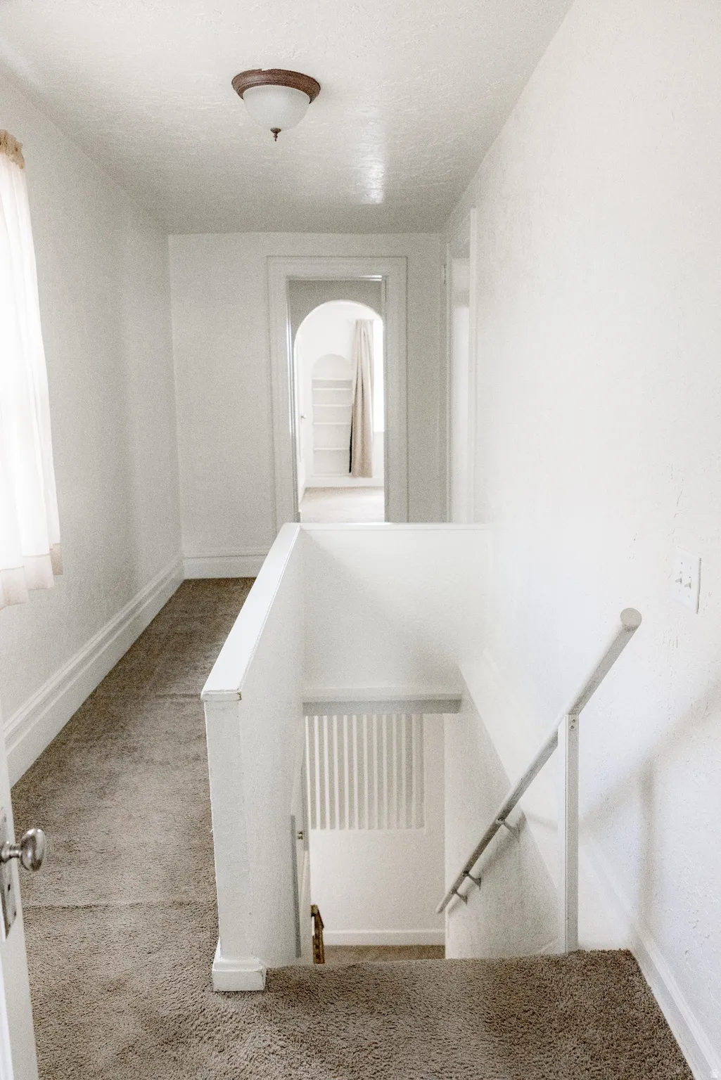 Carpeted hallway with a staircase, white painted walls, and a ceiling-mounted light fixture
