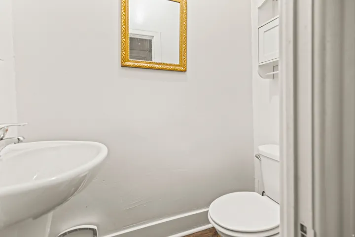Pedestal sink with chrome faucet, gold-framed mirror, white wall-mounted storage, toilet, and wood-finish flooring