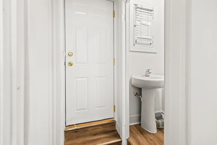 White paneled door with brass hardware, wood-finish flooring, and a white pedestal sink with a window featuring blinds