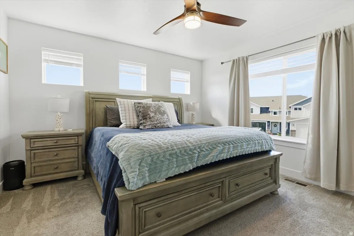 Spacious room featuring neutral carpeting, a large window with roller shades, three transom windows, a ceiling fan with integrated lighting, and white wall finishes