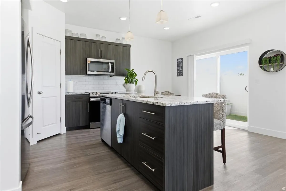 Kitchen featuring dark wood-finish cabinetry, stainless steel appliances, a subway tile backsplash, and a large island with a light-toned granite countertop and integrated sink
