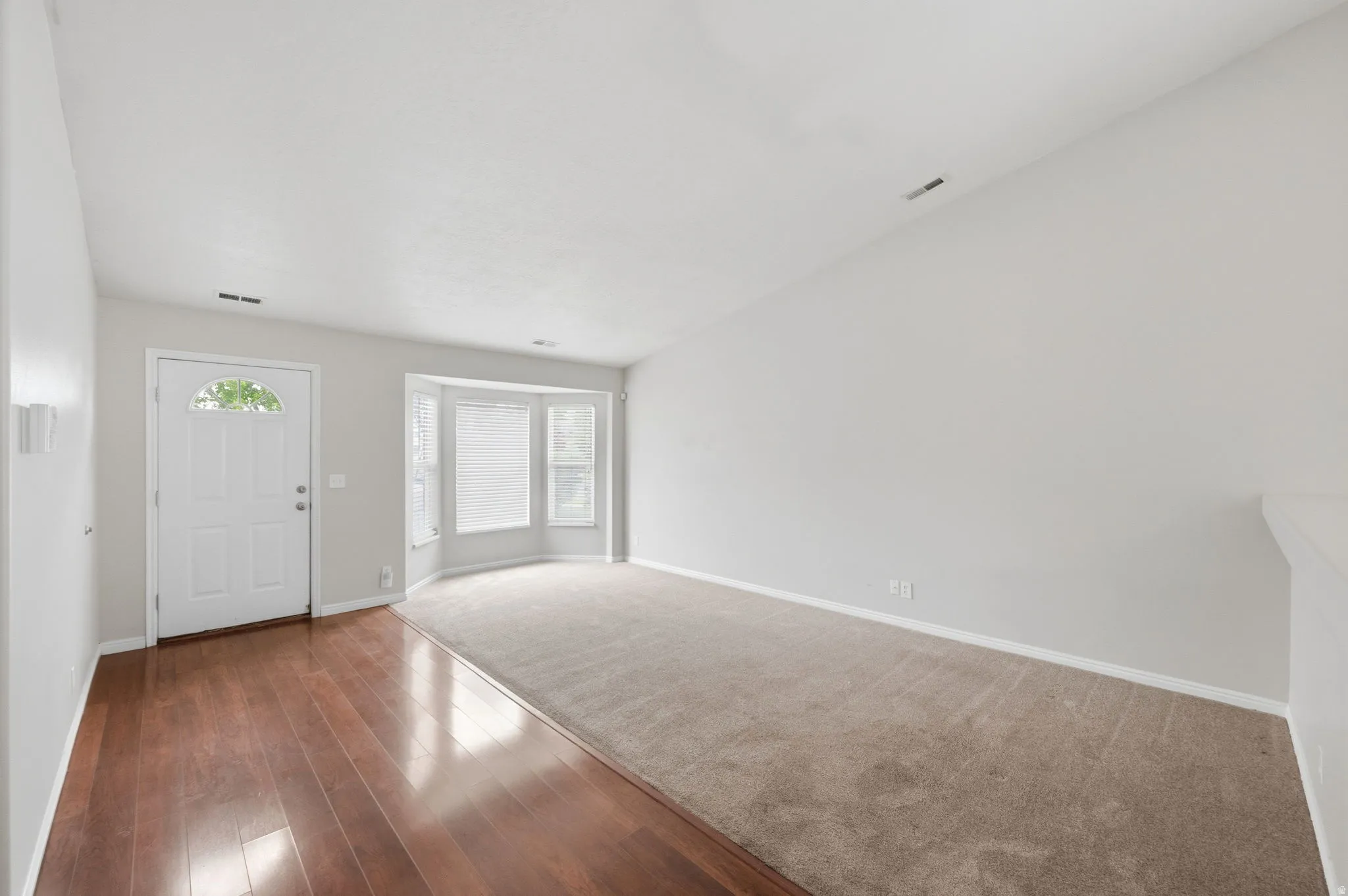 Entryway featuring wood-finish flooring, a white paneled door with an arched window, and a bay window in the carpeted living area