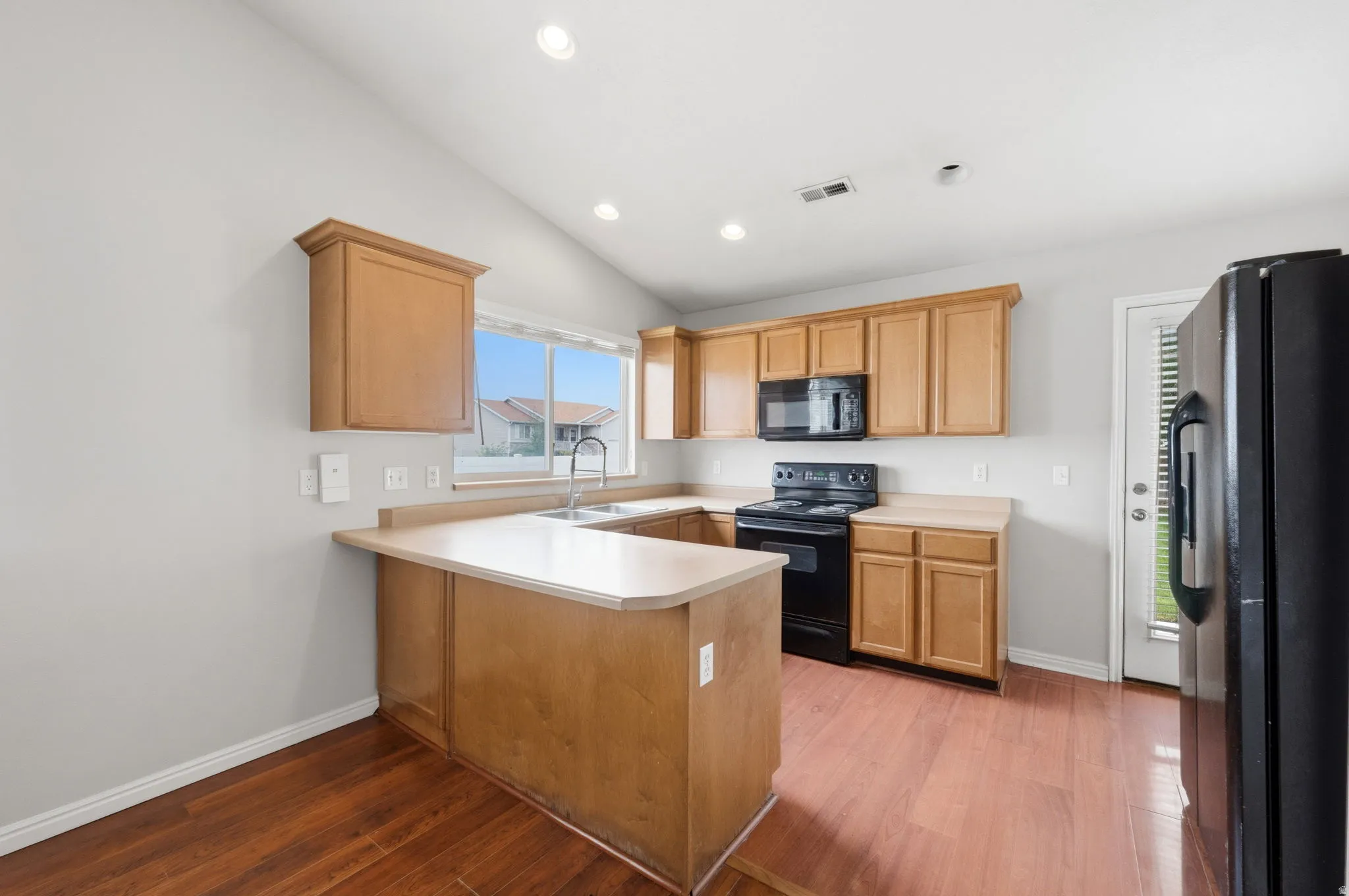 Kitchen featuring wood-finish flooring, light wood cabinetry, light-toned countertops, a stainless steel sink, and black appliances