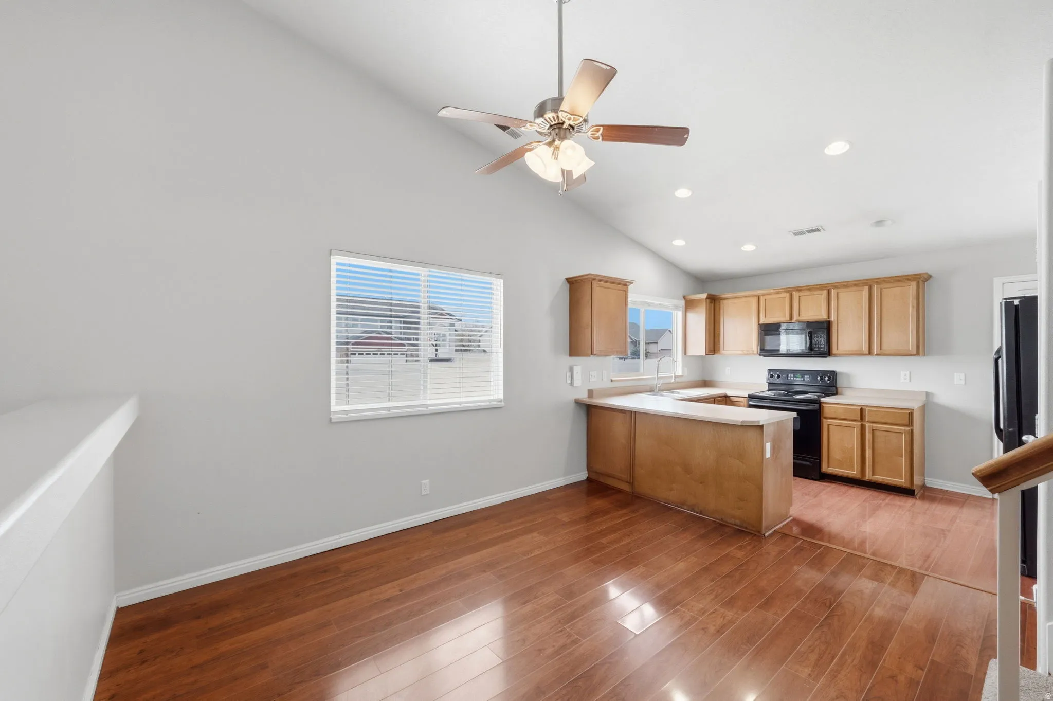 Open-concept living space featuring wood-finish flooring, a vaulted ceiling with a ceiling fan, and a kitchen with warm-toned cabinetry, a black electric range, and a black built-in microwave
