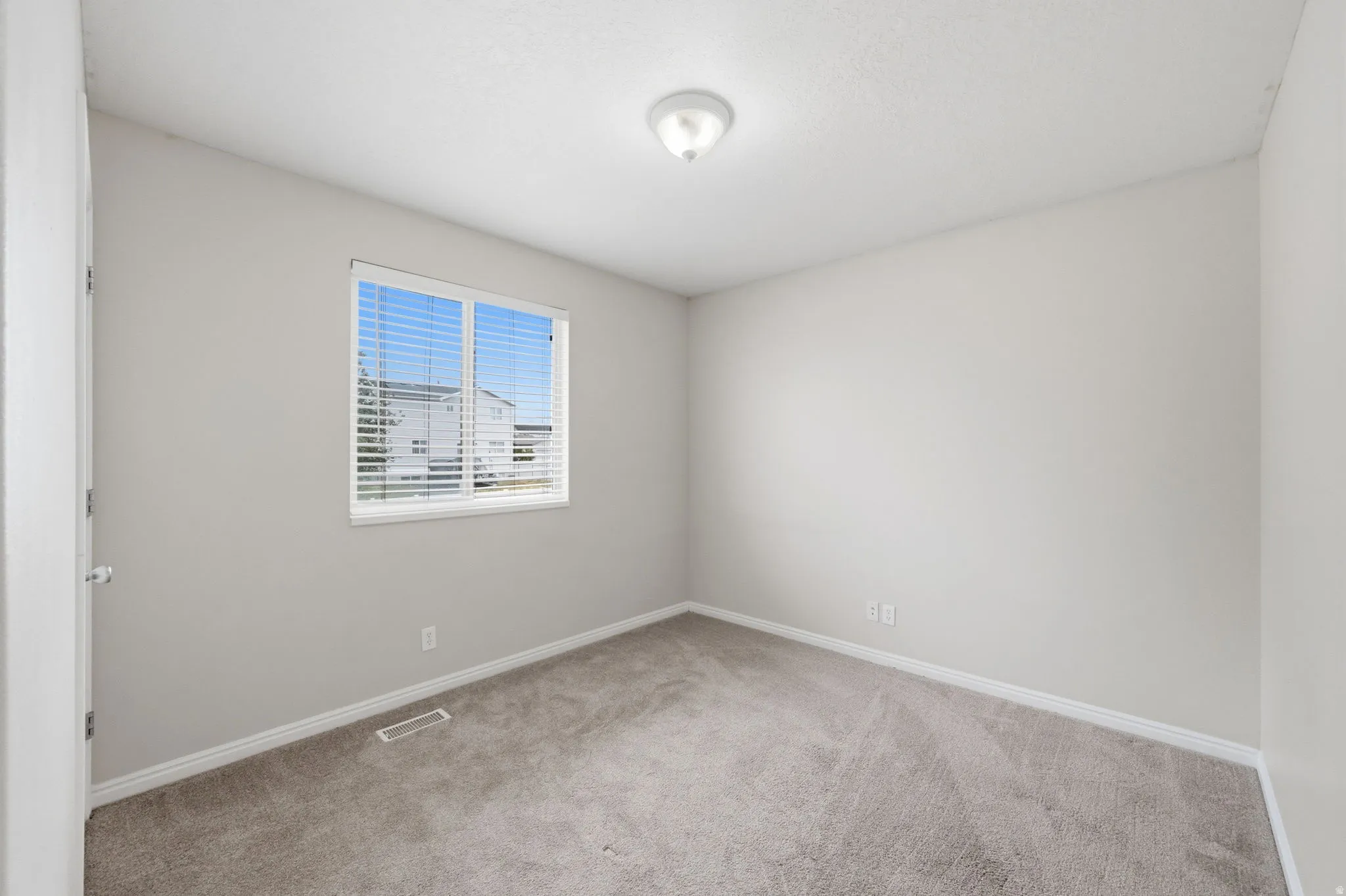 Neutral-toned interior room featuring light gray wall paint, gray carpet, white trim, a single window with horizontal blinds, and a flush-mount ceiling light fixture